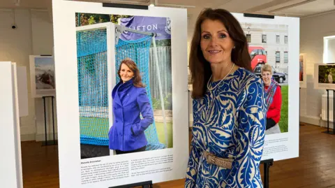 A middle-aged woman with long brown hair and a swirly patterned blue and white dress smiles as she stands beside a photograph of herself with text beneath it at a photography exhibition.