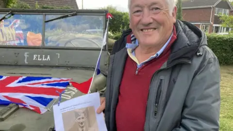 A man wearing a grey coat and red fleece is smiling at the camera and holding a picture of his father. An old green vehicle is behind him.