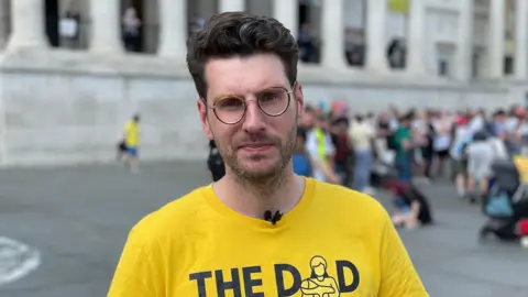 Man with dark hair and glasses wearing a yellow t-shirt with "The Dad Shift" written in black across the front, looking at the camera with a blurred city street behind him.