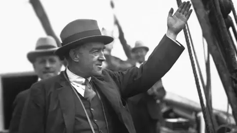 Getty Images A black and white photo of Ernest Shackleton. He is standing on the deck of a ship, wearing a hat, jacket, tie and waistcoat. Two other men, wearing similar attire, are visible in the background.