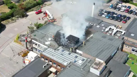 An aerial photograph of Ormiston Cliff Park Primary Academy on Orde Avenue in Gorleston. Fire service members are seeing working on the roof which is charred and has smoke billowing out of it. 