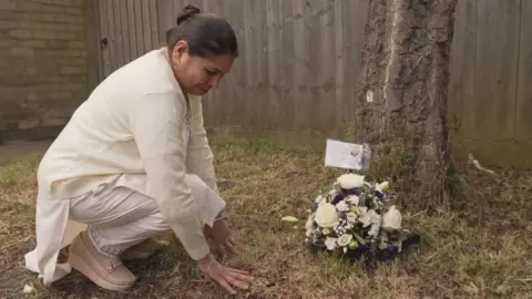 ITV News/PA Sudesh Kumari squats on the lawn of a small garden, next to a tree. Her face is curled up with sorrow. There is a posy of white and purple flowers next to the tree with a note. She wears traditional white Indian clothing.