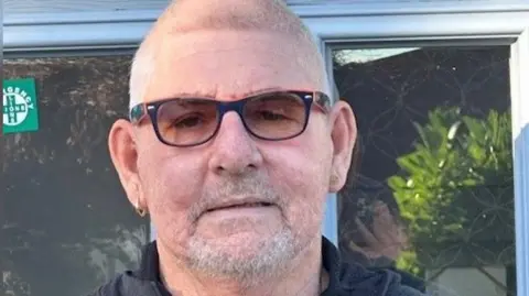 Philip Willis looking straight into the camera, wearing dark framed glasses, facial hair and a gold hoop earring in his left ear and a black jumper