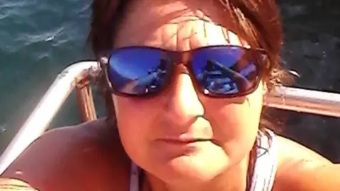 Parker family A woman wearing a white top, dark sunglasses with short brown hair takes a selfie. The sea can be seen behind her