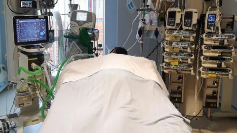 Family photo Eshan Bilal lying comatose in a hospital bed with white sheets surrounded by medical machinery