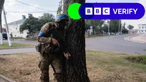 Getty Images A Ukrainian soldier fights in the Kursk region on the Russian border. He is holding a gun and hiding behind a tree, wearing a helmet and military fatigues. 