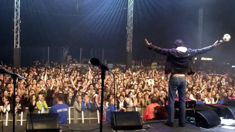 Vernon Kay facing a crowd of people on a stage. He is wearing a black jacket and blue jeans. He has his arms wide. The crowd are all in rapt applause. It is dark. 