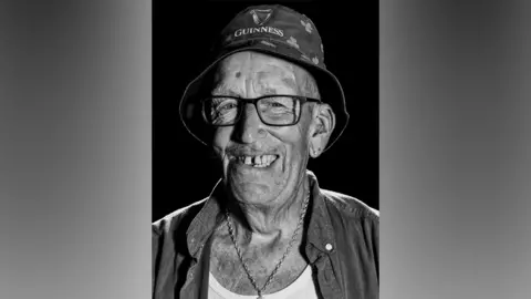 A black-and-white photo of a man smiling into the camera with a black background. He is wearing a bucket hat with the Guinness logo covering his hair and rectangular glasses. He is smiling with his teeth which are large with a gap in the middle. He is also wearing a long chain around his neck and a shirt with a white top underneath. He has two stud earrings in one ear.