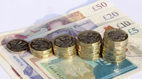 Piles of pound coins which are rising in height from left to right and piled on some UK bank notes