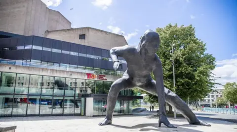 The front of the theatre building. It is a large glass and concrete modern building. A large statue showing a woman with one hand touching the ground is in front of the entrance. The 7-metre tall bronze is the UK's largest bronze sculpture, created by artist Joseph Hillier.