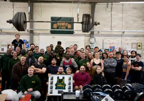 Charlotte Macaulay Two dozen people pose for a picture taken in the gym with a large weight above them