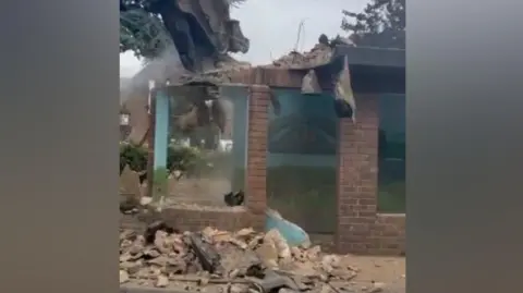 An excavator with a large pneumatic drill attachment breaks up a brick bus shelter. It is working on the roof and a mural on a back wall can just be seen through the dust.