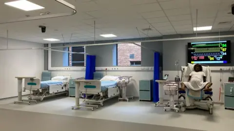 A wide shot of a mock-maternity ward at York St John's University - complete with hospital beds, and the Mama Anne manikin in a bed. On a TV screen, a heart rate monitor can be seen.
