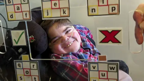A young man in a chair smiling at the camera, as seen through a transparent board with multiple coloured letters and symbols. The man has an oxygen line attached to his face.