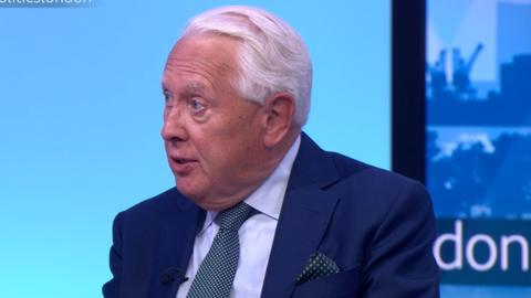 Sir Bob Neill wearing a navy blue suit sits in front of a blue backdrop