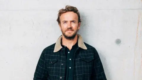 Matt Porteous Nick Hounsfield standing against a white concrete wall. He is wearing a dark buttoned shirt and a checked patterned jacket with a fluffy collar. He has blue eyes, short brown hair, a full brown beard, and is smiling at the camera.