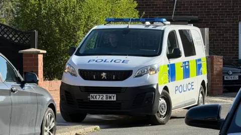 Claudia Robinson/BBC A white police van with yellow and blue squares on the side of the van. 'Police' is written across the front and the side in blue writing. It is half parked on the curb and there is a man sat inside the car.