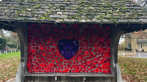 Holt Residents A bench with wooden shelter and back, a sea of red poppies with a purple heart with purple poppies in the middle