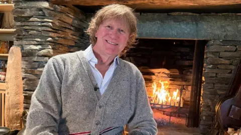 Sir Chris Evans has a mop of blonde hair and is wearing a grey cardigan sat next to a large roaring fire