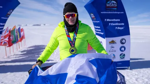 World Marathon Challenge Bobby pictured wearing a florescent yellow top, a black woolen snood and sunglasses. He is wearing a medal and holding a saltire flag with a finish line in the background.
