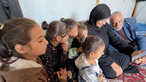 Huda Abu Jazar, wearing a black dress and headgear, holds a mobile phone, as she sits on a carpet with a man on her left, wearing blue trousers, dark T-shirt and a light blue blazer, and four children to her right - all watching the mobile phone