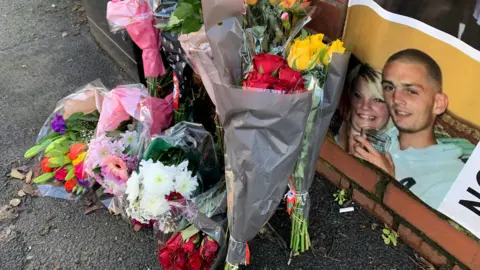 Emma Daniels Floral tributes on a pavement next to a picture of Matthew Schofield on the wall