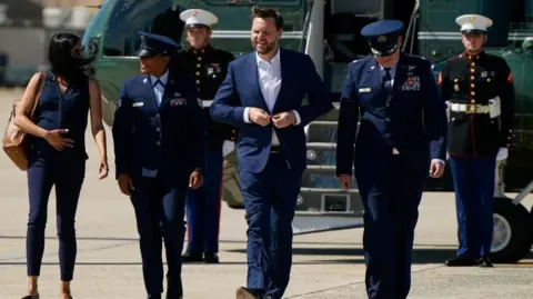 US Vice President JD Vance disembarks Marine Two flanked by soldiers and his wife Usha Vanceas he arrives at Joint Base Andrews, Maryland. He is wearing a blue suit and is buttoning his jacket. 