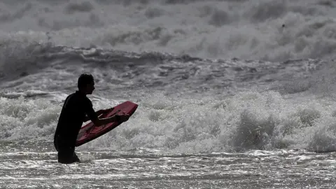 A man with a small red surf board is standing in rough grey seas