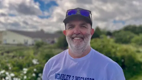 Ant Saddington/BBC Damien Johnston standing outside with shrubs behind him. He is wearing a baseball cap, with dark glasses propped on its brim and a white T-shirt with Wembley 2 Wigmore written on it. He has a short cropped white beard and is smiling. 