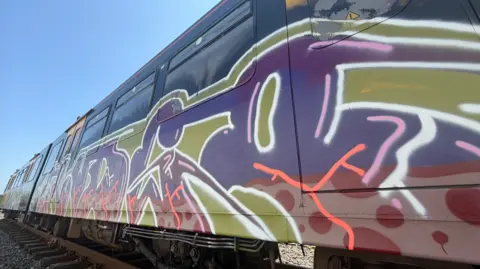 BBC/Jim Scott Graffiti of green, purple and red colourings has replaced the traditional grey, black and yellow colour scheme of the Metro car. There are also black spray marks covering the windows.