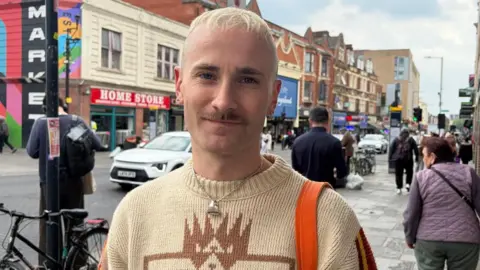 Kris standing on a Dalston street, wearing a patterned cream jumper and carrying an orange bag.