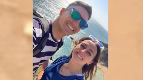 Family handout Adam Harrison and his girlfriend Chelsea smile as Adam takes a selfie. He has blue sunglasses on along with a grey backpack and blue and white striped T-shirt. Chelsea has white and blue sunglasses and a blue shirt on. They are standing in front of a beach and the sea.