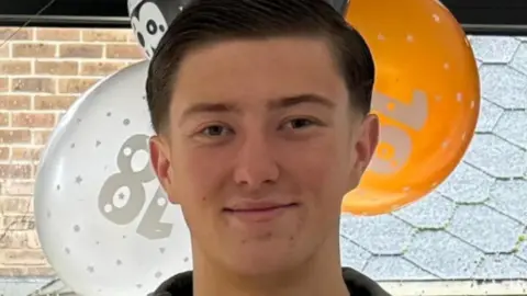 Police handout An 18-year-old man standing in front of orange, white and black balloons by a window. He has short dark hair and is smiling and is wearing a blue jacket and a black top underneath.