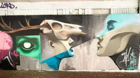 James Thompson/Wall Games A street art mural featuring two people facing each other and an erratic contrast of colours. One is wearing a deer skull on their head.