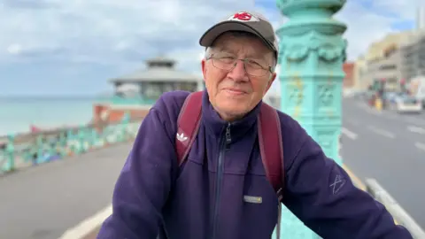 An elderly man with glasses. He is wearing a purple fleece and a grey baseball cap. He is standing on Brighton seafront, with a road behind him to his right, and the sea behind him to his left