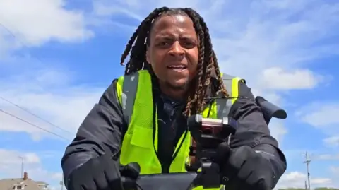 The streamer Isaiah Thomas, known as hmblzayy, walking while wearing a high-vis jacket and pushing a cart in front of him on a sunny day.