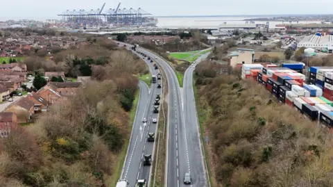 Shaun Whitmore/BBC A drone image shows a dual carriageway with tractors on the left hand carriageway heading towards Felixstowe, whose cranes are visible in the distance. The tractors are driving in convoy behind the other in the fast lane. Housing is visible next to the road on the left, with containers stacked up on the right.