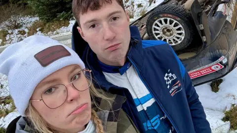 Hollie McRae Hollie McRae and her co-driver, Jon Armstrong, with sad expressions standing in front of the upturned rally car