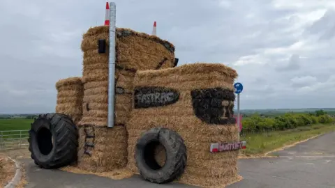 Huntingdon Young Farmers A straw bale sculpture showing a tractor. It has very fat black tyres resting on its side and two orange and white cones resting on its top. Its cab has a window highlighted in black and beneath it is a licence plate saying HuntsYF in pink. It is in a farm gate.