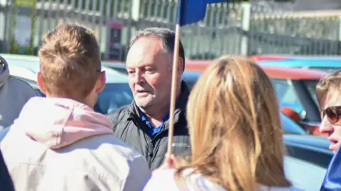 Richard Griffin A man with short grey hair who is in a black coat who is surrounded by people on a picket line. There are several parked cars behind them.