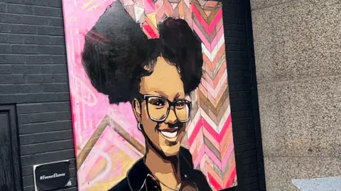 Mural Elianne Andam telah dilukis di dinding di Croydon di London selatan. Di dalamnya dia tersenyum dan memakai kacamata. Rambutnya terangkat. Ini menunjukkan kepala dan bahunya dan latar belakangnya adalah desain merah muda dengan pola geometris