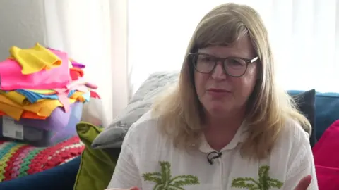 Blonde woman being interviewed indoors with stacks of brightly coloured tea towels piled behind on a sofa.
