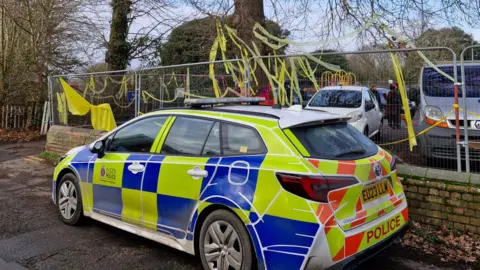 James Fell/BBC A police car parked in front of an oak tree, which has yellow ribbons attached to its branches. The tree is being held behind heras fencing. Campaigners are stood in the background.