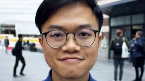 University of Reading Headshot of Dr Kiwi Tang standing in a street. He is wearing round glasses and has short black hair.