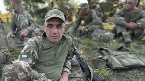 Rostyslav in military uniform with a cap on, leaning over as he sits on a field surrounded by other Ukrainian recruits