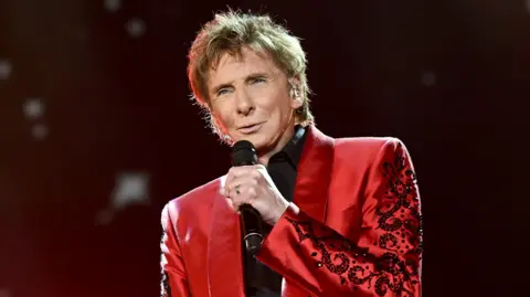 James Watkins/BBC Barry Manilow sings into a microphone on a stage. He wears a red velvet suit with a black shirt underneath. He has blonde hair.