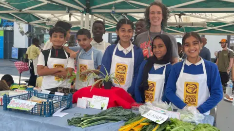 Children gathered with one of their teachers at their stall in the market. On the table are green vegetables including lettuce and chard, as well as potted spider plants and a box full of potatoes.