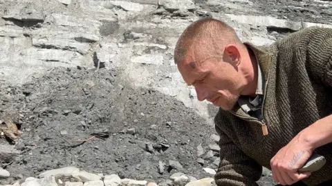 Casey is on a rocky beach, he is seen in profile from his left, bending down and looking at something on the ground out of frame. In the background are the bottoms of cliffs and loose rock. 