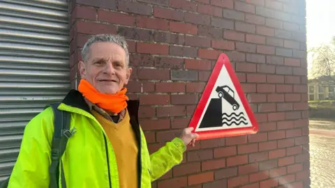 A man with short grey hair wearing a yellow high viz coat, yellow jumper and orange neck protector points at a triangular red sign warning drivers not to drive into the river. 
