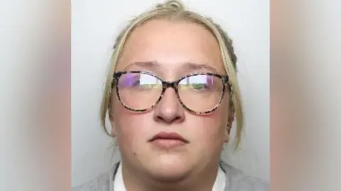 A mugshot of a woman with blond hair in a ponytail, wearing a grey jumper, and tortoiseshell glasses.